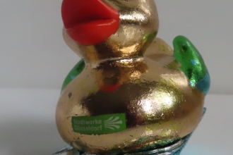 Düsseldorfer Ente 2025, Plastikfigur auf Holzwippe, Metallblatt vergoldet mit Glitzersteinen und Spiegel beklebt.